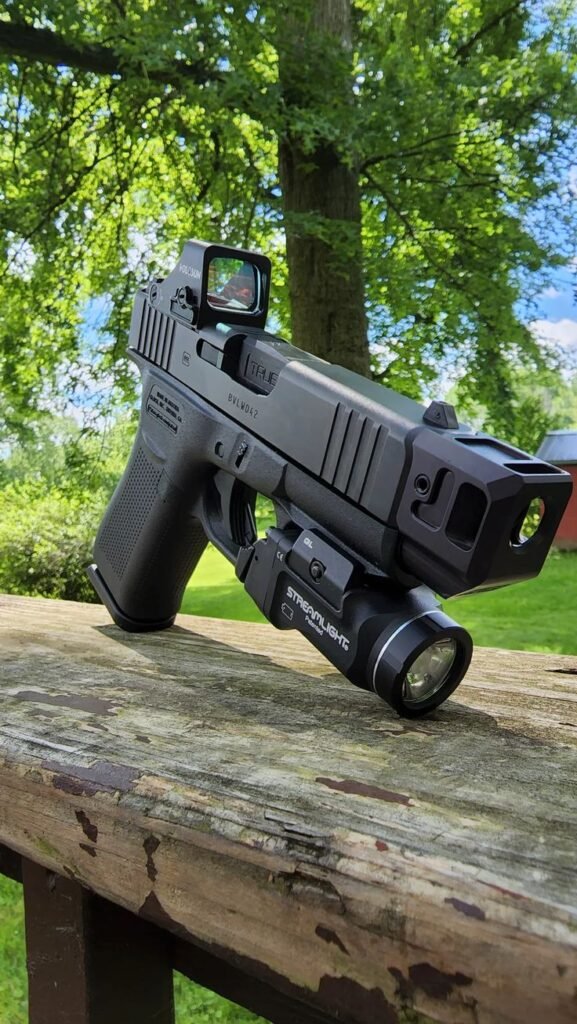 Custom Glock pistol with a red dot sight, compensator, and mounted flashlight resting on a wooden surface with greenery in the background.